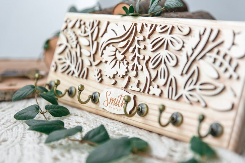 May include: Wooden key holder with a floral design and five hooks. The key holder has a sign that reads "Smith Family".
