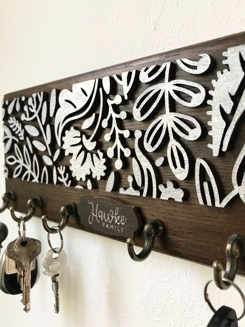 May include: A wooden key holder with a floral design and a metal plaque that reads "Hawke Family". The key holder has five hooks for hanging keys.