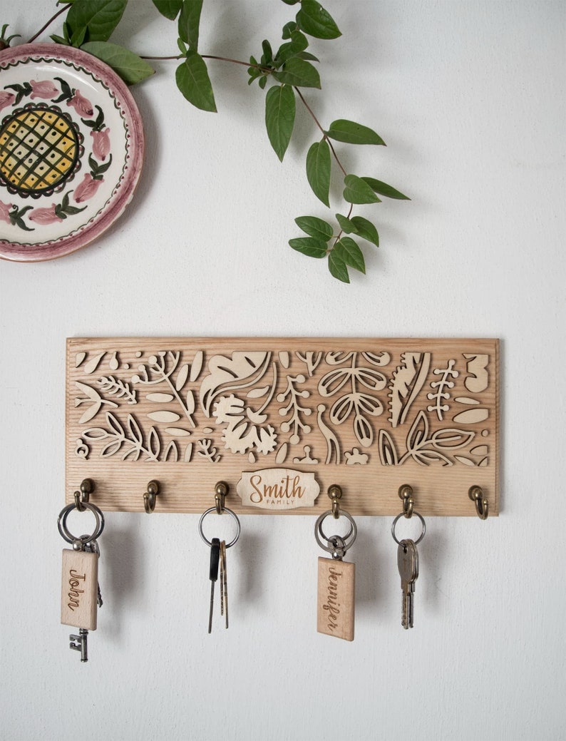 May include: A wooden key holder with a floral design and five hooks. The key holder has a small plaque that says "Smith Family". There are three keys with personalised tags that say "John", "Jennifer", and "Trevor".