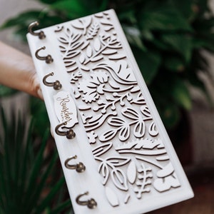 May include: White wooden key holder with a floral design and five hooks. The key holder has a wooden plaque that says "Rauchcher Family".