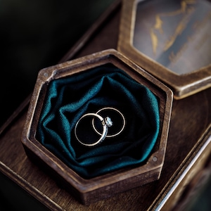 May include: Two gold wedding rings rest in a wooden hexagonal box lined with dark teal fabric. One ring features a small diamond. The box is open, revealing the rings, and sits on a larger wooden surface.