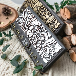 May include: Two wooden key holders with a floral design. The key holder on the left has a white floral design on a dark brown background. The key holder on the right has a gold floral design on a dark brown background. Both key holders have five hooks.