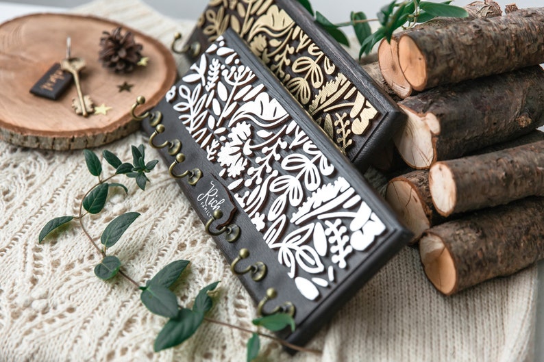 May include: Two wooden key holders with a floral design. The key holder on the left has a white floral design on a dark brown background. The key holder on the right has a gold floral design on a dark brown background. Both key holders have five hooks.