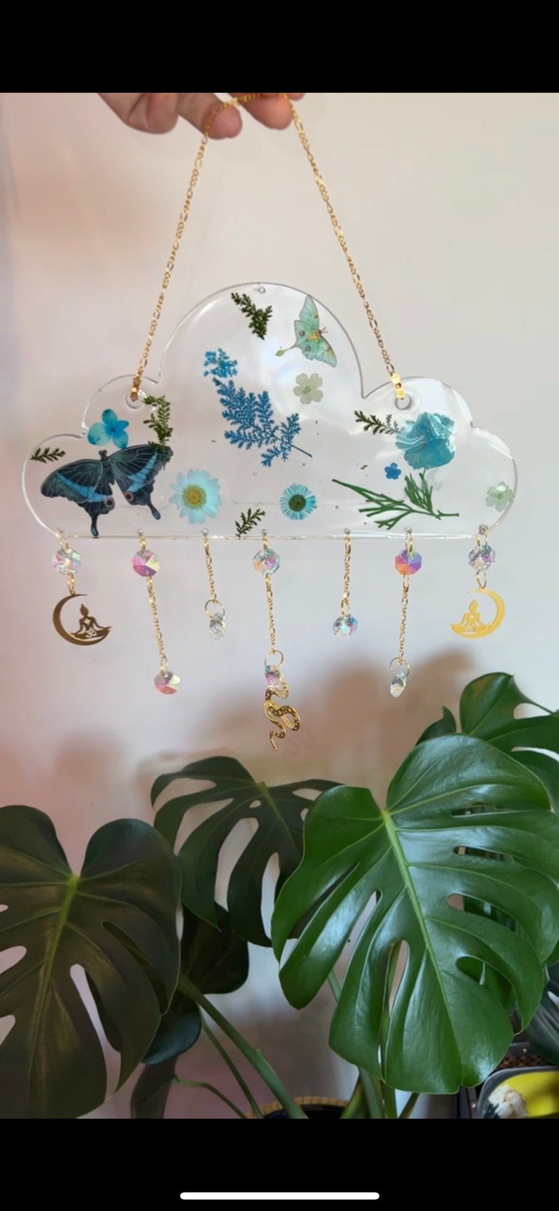 May include: A clear resin cloud-shaped ornament with pressed flowers and butterflies, hanging from a gold chain with crystal and gold charms. The charms include a crescent moon with a meditating figure, a snake, and a crystal.