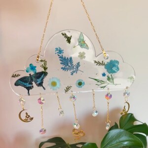 May include: A clear resin cloud-shaped wall hanging with pressed flowers and butterflies. The hanging has a gold chain and multiple crystal and gold charms dangling from it.