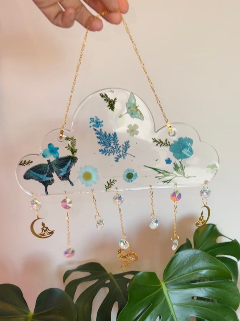 May include: A clear resin cloud-shaped wall hanging with pressed flowers and butterflies. The hanging has a gold chain and multiple crystal and gold charms dangling from it.