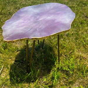 May include: A purple and white resin table top with gold accents and gold metal hairpin legs. The table is sitting on a green grassy surface.