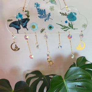 May include: A clear resin cloud-shaped ornament with pressed flowers and butterflies, hanging from a gold chain with dangling crystals and gold charms of a crescent moon and a snake.