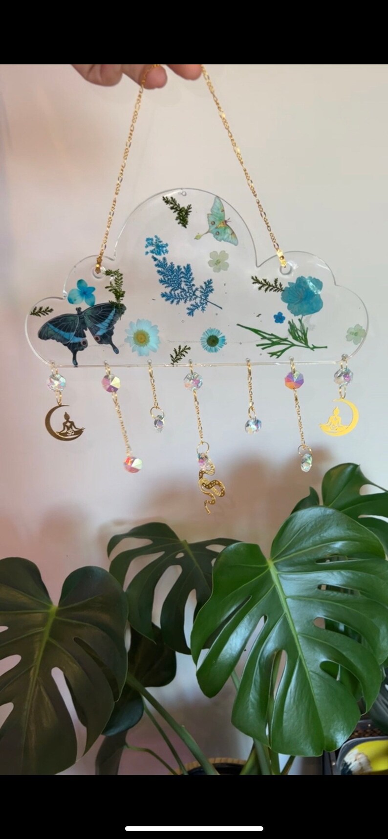May include: A clear resin cloud-shaped ornament with pressed flowers and butterflies, hanging from a gold chain with dangling crystals and gold charms of a crescent moon and a snake.