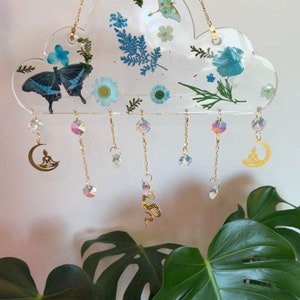 May include: A clear resin cloud-shaped wind chime with pressed flowers and butterflies. The chime has gold chains with crystal beads and charms, including a crescent moon with a meditating figure and a snake.