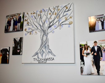 May include: A white canvas with a grey and blue painted tree with finger prints on the branches. The canvas has the text "TYLER + KARISSA 10.08.17" at the bottom. The canvas is surrounded by framed photos of a wedding party, a couple, and two dogs.