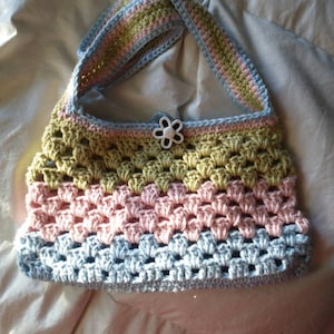 May include: A crocheted tote bag with a light blue, pink, and green striped strap. The bag is made with a simple crochet stitch and has a light blue, pink, and green color scheme. A white flower button is attached to the front of the bag.