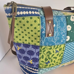 Puede incluir: Bolso tote patchwork con cuadrados estampados en tonos azules, verdes y blancos. Cuenta con asas de cuero marrón y cierre de cremallera. Los estampados incluyen lunares, diseños florales y motivos de árboles y pájaros.
