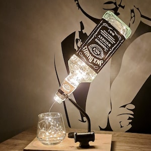 Può includere: Una lampada a forma di bottiglia di whisky Jack Daniel's con un bicchiere e luci a catena. La bottiglia è trasparente e ha una superficie testurizzata. La lampada è su una base di legno.