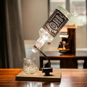 Peut inclure: Une bouteille de whisky Jack Daniel's avec des lumières de fée à l'intérieur est montée sur une base en bois et se verse dans un verre avec des lumières de fée. La bouteille est inclinée et le verre est posé sur la base.