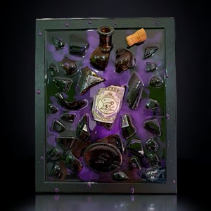 Può includere: Un'opera d'arte incorniciata che presenta una bottiglia di vetro rotta su uno sfondo di resina viola. La bottiglia è piena di piccoli pezzi di vetro nero e un tappo di sughero. L'opera è firmata "M. Butler" nell'angolo in basso a destra.