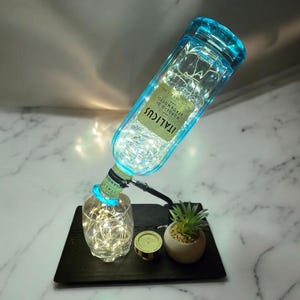 Op de afbeelding: Een hergebruikte fles met een blauwe dop en het woord "ITALICUS" op het etiket, verlicht door feeërieke lichtjes. De fles is verbonden met een klein glas en een kleine potplant op een zwart dienblad.
