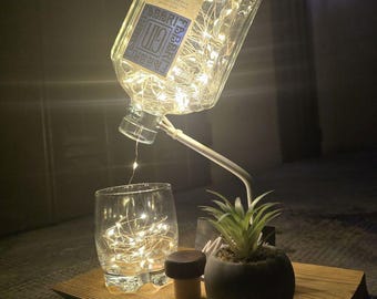 May include: A clear glass bottle with a label that reads "DRY GIN" and "FABER'S FARM" is filled with white fairy lights. The bottle is suspended above a glass with fairy lights, a small potted plant, and a cork stopper on a wooden tray.
