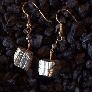 May include: A pair of gold earrings with a cube-shaped pyrite stone hanging from each. The stones are a silvery gray color with a metallic sheen.