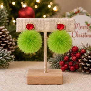 May include: A pair of earrings featuring a bright green fluffy pom-pom attached to a gold-tone stud with a red heart-shaped gemstone. The earrings are displayed on a beige T-shaped stand, showcasing the vibrant green texture and red accents.