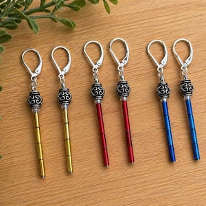 Op de afbeelding: Zes paar oorhangers met zilverkleurige oorhaaksluitingen. De oorbellen hebben een decoratieve zilveren kraal en zijn verkrijgbaar in goud, rood en blauw. De oorbellen worden gepresenteerd op een houten oppervlak.