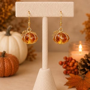 May include: A pair of gold-toned dangle earrings featuring faceted amber-colored glass beads shaped like pumpkins. Each earring is suspended from a gold-toned hook and includes a small wire coil detail at the top, resembling a pumpkin stem.