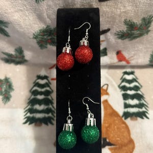 May include: Two pairs of Christmas ornament earrings. One pair features red glitter ornaments, and the other pair has green glitter ornaments. Both pairs have silver-colored hooks and caps. The earrings are displayed on a black velvet stand against a festive background.