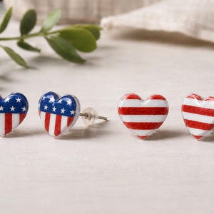 May include: Heart-shaped stud earrings with patriotic designs. Two pairs of earrings are visible: one pair features the American flag, and the other pair has red and white stripes. The earrings are on a light-colored surface.