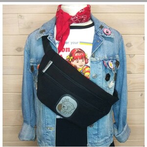 Puede incluir: Riñonera negra con un parche de dibujos animados, usada sobre una chaqueta vaquera y una camiseta blanca. La riñonera tiene un bolsillo con cremallera y un pequeño logotipo. Un pañuelo rojo está atado al cuello.