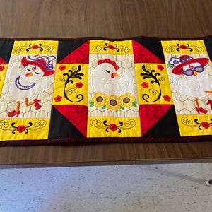 May include: A decorative table runner featuring embroidered chickens wearing hats and sunglasses. The runner has a black border, red and yellow panels, and floral accents. The chickens are white with red combs and are wearing hats and sunglasses.