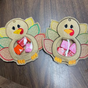 May include: Two felt turkey-shaped pouches with embroidered details. Each pouch has a clear plastic window to hold small treats. The turkeys are decorated with brown, orange, yellow, and green felt.