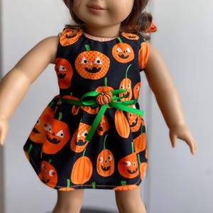 May include: A doll wearing a black Halloween dress with orange jack-o'-lanterns. The dress has a green ribbon belt with a pumpkin accent. The doll has brown hair and blue eyes. The dress is sleeveless.