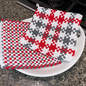 May include: Two red and gray woven pot holders on a white plate. The pot holders are made of a thick, textured fabric and have a checkered pattern.