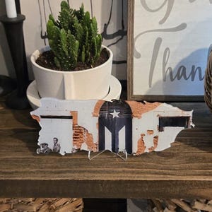 May include: A wooden shelf unit with three shelves, each holding various items. The top shelf has a sign that reads "In all things give thanks", a small potted succulent, two black candle holders with white candles, and a gray ceramic vase. The middle shelf has a wooden cutout of the state of Texas, a woven basket, and a book titled "Madrid London". The bottom shelf has three woven baskets.