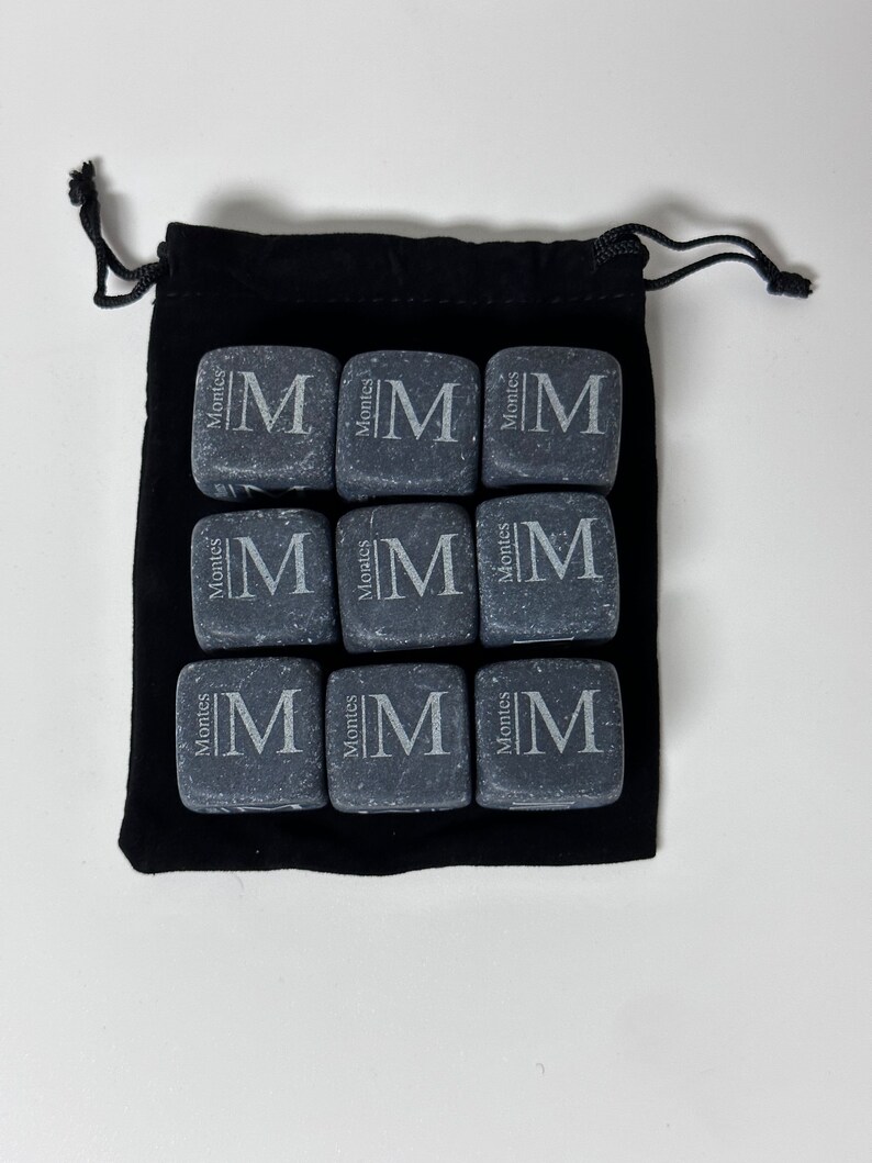 May include: Nine grey stone cubes with the letter "M" and the word "Montes" engraved on each cube. The cubes are in a black velvet drawstring bag.