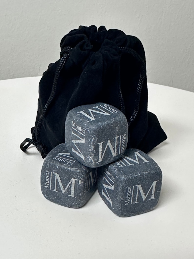 May include: Four grey stone ice cubes with the word "Montes" and the letter "M" engraved on each. The ice cubes are in a black velvet drawstring bag.