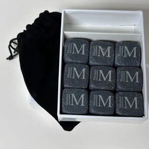 May include: A set of nine grey stone whiskey chilling stones in a white box with a black velvet bag. Each stone is engraved with the letter "M" and the word "Montes".