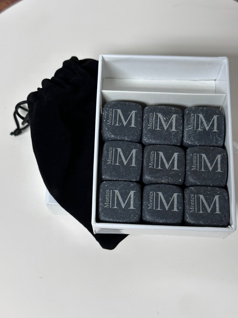 May include: A set of nine grey stone whiskey chilling stones in a white box with a black velvet bag. Each stone is engraved with the letter "M" and the word "Montes".