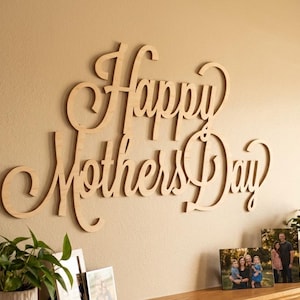 May include: Wooden wall art spelling "Happy Mothers Day" in a cursive font. The sign is mounted on a beige wall above a shelf with framed photos and potted plants. A decorative piece for Mother's Day.