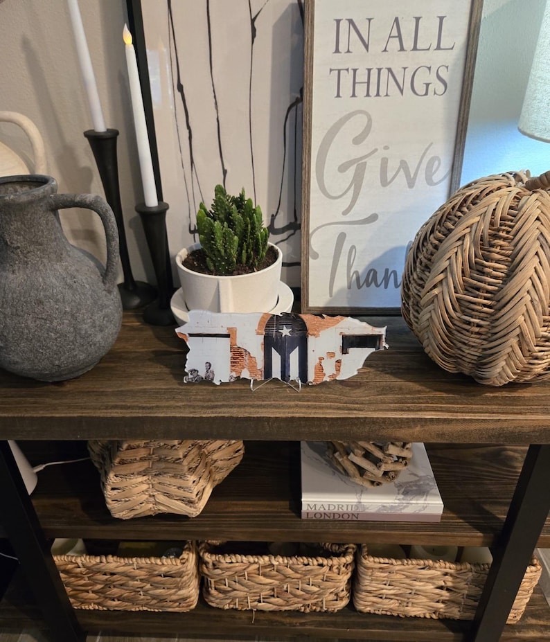May include: A wooden shelf unit with three shelves, each holding various items. The top shelf has a sign that reads "In all things give thanks", a small potted succulent, two black candle holders with white candles, and a gray ceramic vase. The middle shelf has a wooden cutout of the state of Texas, a woven basket, and a book titled "Madrid London". The bottom shelf has three woven baskets.