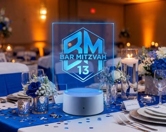 May include: A clear acrylic sign with a blue glowing hexagonal logo featuring the letters RM, the text BAR MITZVAH, and the number 13. The sign is mounted on a circular white base that illuminates the design with a bright blue light.
