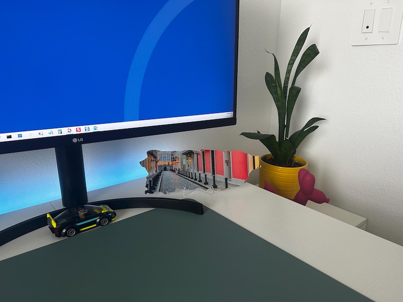 May include: A computer monitor with a blue screen is on a desk with a small toy car, a plant in a yellow pot, and a pink figurine.