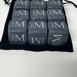 May include: Nine grey stone cubes with the letter "M" engraved on each side. The cubes are arranged in a 3x3 grid and are sitting on a black velvet pouch.