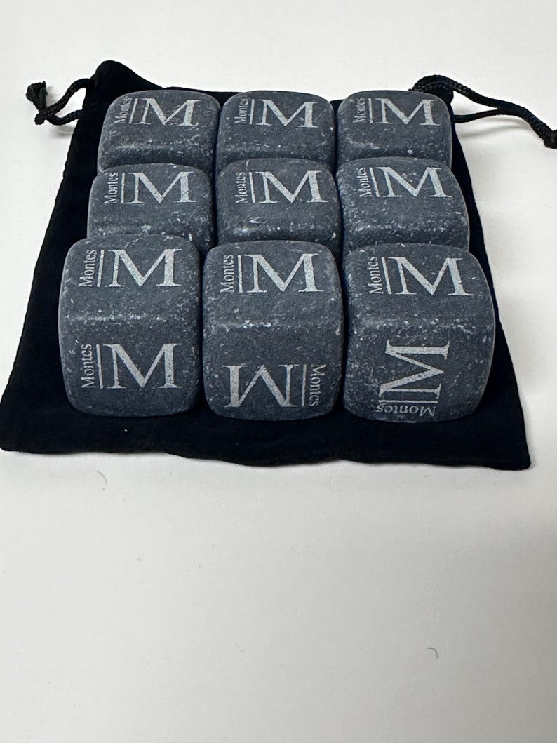 May include: Nine grey stone cubes with the letter "M" engraved on each side. The cubes are arranged in a 3x3 grid and are sitting on a black velvet pouch.