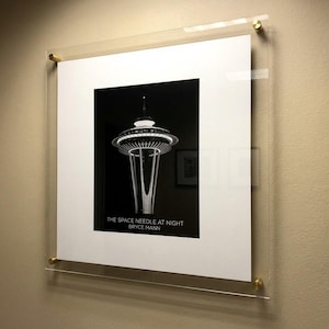 May include: Framed black and white print of the Space Needle at night, mounted on a wall. The print is encased in a clear acrylic frame with gold-colored hardware. The text "THE SPACE NEEDLE AT NIGHT BRYCE MANN" is visible.
