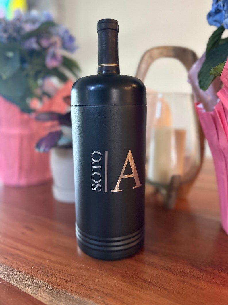 May include: Black wine bottle-shaped container with the name "SOTO" and the letter "A" in a gold color. The container has a black top and a gold band around the neck. The container is sitting on a wooden surface.