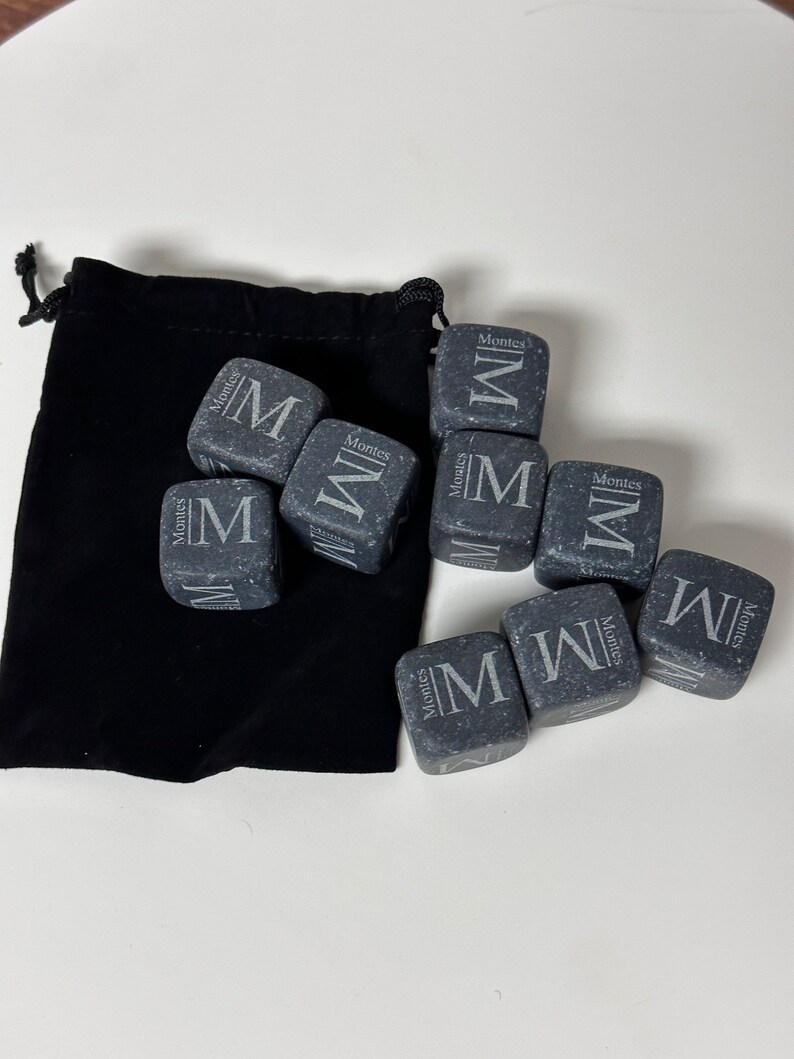 May include: A set of nine grey stone whiskey chilling cubes with the word "Montes" and the letter "M" engraved on each cube. The cubes are in a black velvet pouch.
