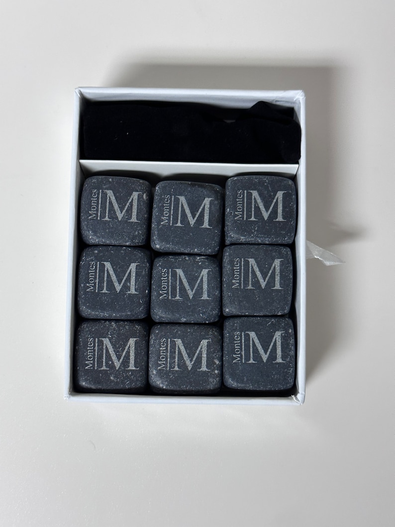 May include: A white box with a black velvet lining contains nine grey stone coasters. Each coaster has the letter "M" and the word "Montes" engraved on it.