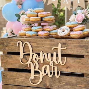 May include: A wooden donut bar display with a variety of donuts with pink and blue frosting. The donuts are arranged on wooden trays and a tiered stand. The words "Donut Bar" are cut out of wood and displayed on the front of the bar.
