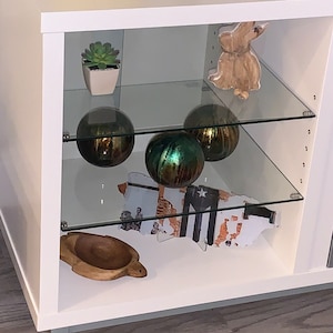May include: White cabinet with glass shelves displaying decorative items. The top shelf has a small potted succulent plant and a wooden rabbit figurine. The middle shelf has three glass orbs, two are gold and one is green. The bottom shelf has a wooden turtle and a framed print of a map.